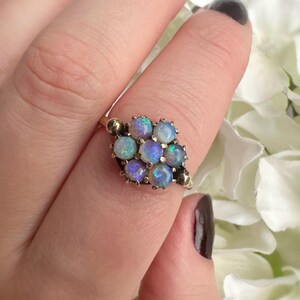 May include: A gold ring featuring a cluster of nine opals. The opals are round and display a range of colors, including blue, purple, and white. The ring has a vintage design.