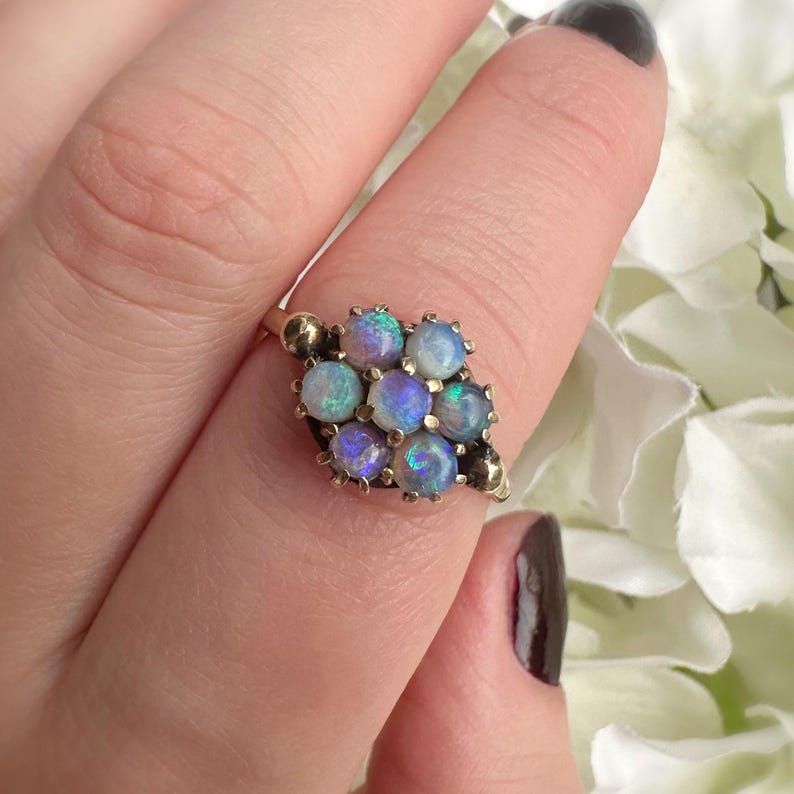 May include: A gold ring featuring a cluster of nine opals. The opals are round and display a range of colors, including blue, purple, and white. The ring has a vintage design.