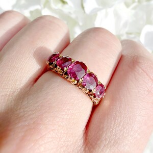 May include: A gold ring featuring five oval-cut, deep pinkish-red gemstones arranged in a row. The metal band displays intricate, decorative metalwork between each stone, creating a textured setting that secures the vibrant, faceted gems.