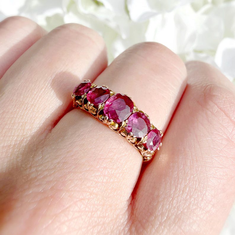 May include: A gold ring featuring five oval-cut, deep pinkish-red gemstones arranged in a row. The metal band displays intricate, decorative metalwork between each stone, creating a textured setting that secures the vibrant, faceted gems.