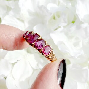 May include: A gold ring featuring four oval-cut deep pinkish-red gemstones arranged in a row. The metal band displays intricate decorative metalwork along the sides, supporting the four faceted stones set in a horizontal line across the finger.