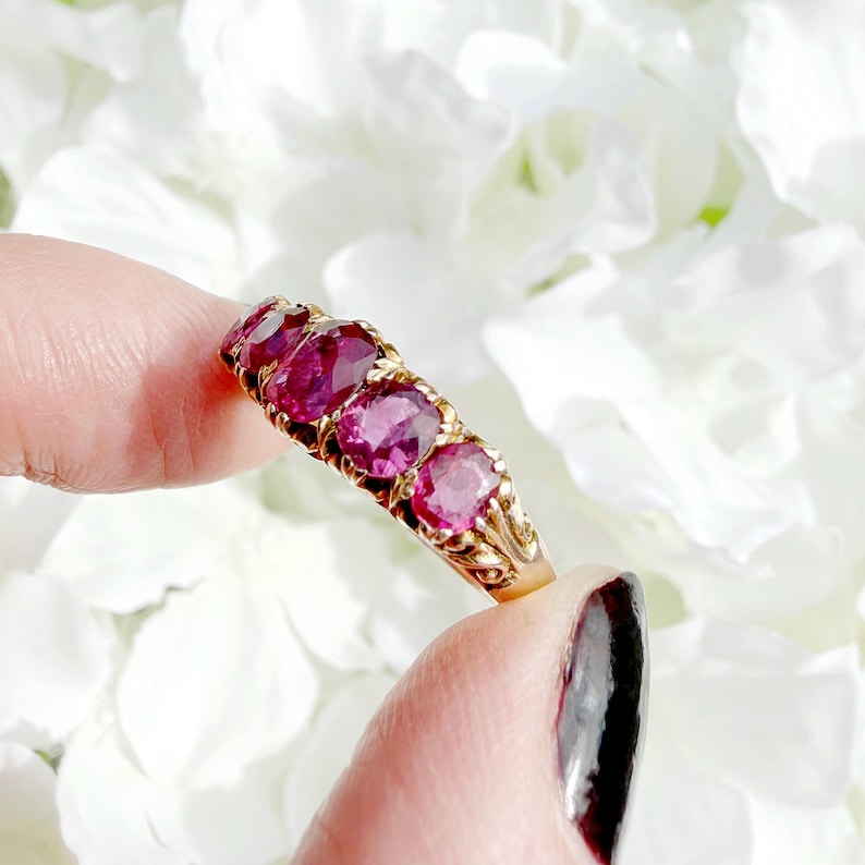 May include: A gold ring featuring four oval-cut deep pinkish-red gemstones arranged in a row. The metal band displays intricate decorative metalwork along the sides, supporting the four faceted stones set in a horizontal line across the finger.