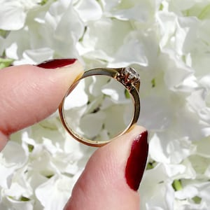 May include: A gold ring with a small, clear gemstone held between fingers with red nail polish. The ring's band is thin and appears to be made of gold. The background is a soft focus of white flowers.