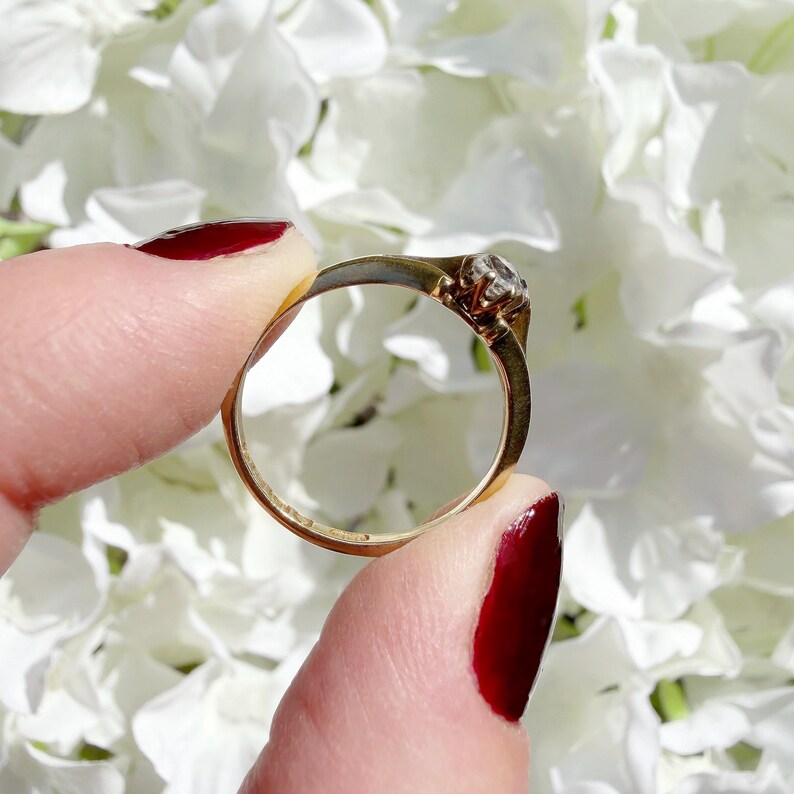 May include: A gold ring with a small, clear gemstone held between fingers with red nail polish. The ring's band is thin and appears to be made of gold. The background is a soft focus of white flowers.