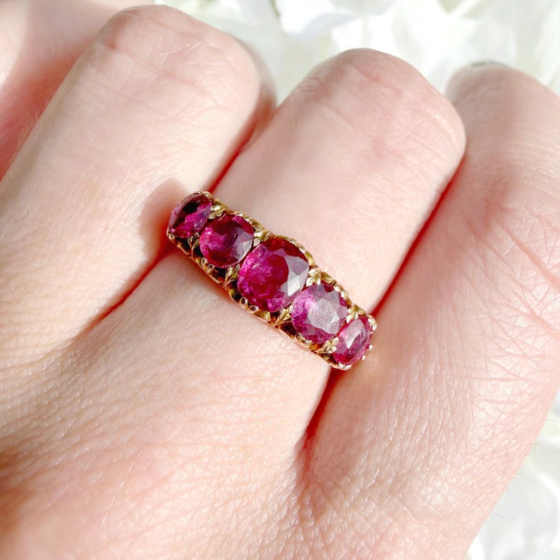 May include: A gold band ring featuring five cushion-cut, deep pinkish-red gemstones arranged in a row. The metal setting has a textured, decorative finish surrounding each stone, creating a continuous, ornate design across the finger.