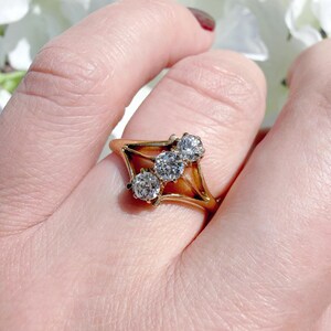 May include: A gold ring featuring three clear, round gemstones set in a geometric design. The ring has a delicate, openwork frame and is shown in a close-up view.