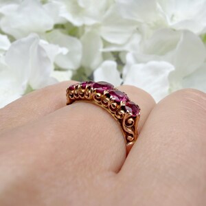 May include: A gold ring featuring a row of five round, deep pinkish-red gemstones set in a decorative, ornate metal band with intricate scrollwork detailing along the sides of the setting, showcasing a classic and detailed craftsmanship.