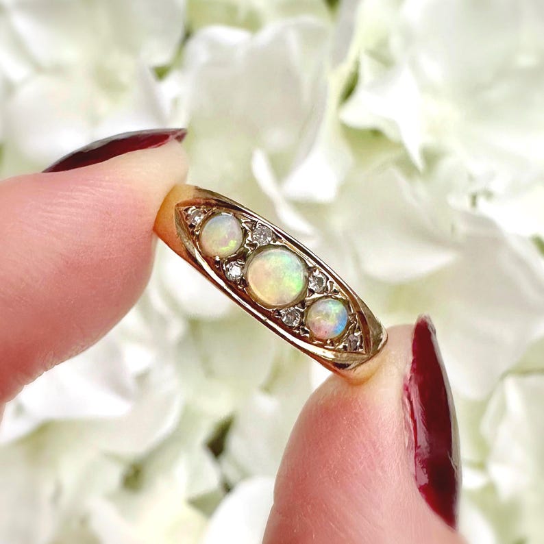 Op de afbeelding: Een gouden ring met drie ovale opalen en kleine diamanten. De opalen vertonen een kleureffect. De ring wordt tussen vingers met rode nagellak gehouden.