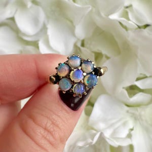 May include: A cluster ring featuring a collection of opals in a gold setting. The opals display a range of colors, including blue, pink, and yellow. The ring is held by a person with a dark manicure, set against a backdrop of white flowers.