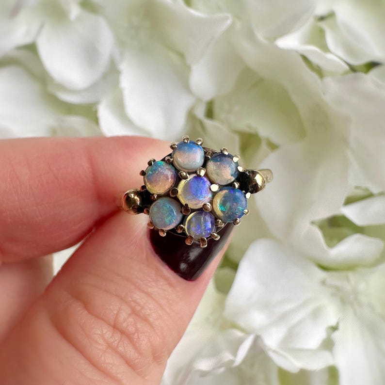 May include: A cluster ring featuring a collection of opals in a gold setting. The opals display a range of colors, including blue, pink, and yellow. The ring is held by a person with a dark manicure, set against a backdrop of white flowers.