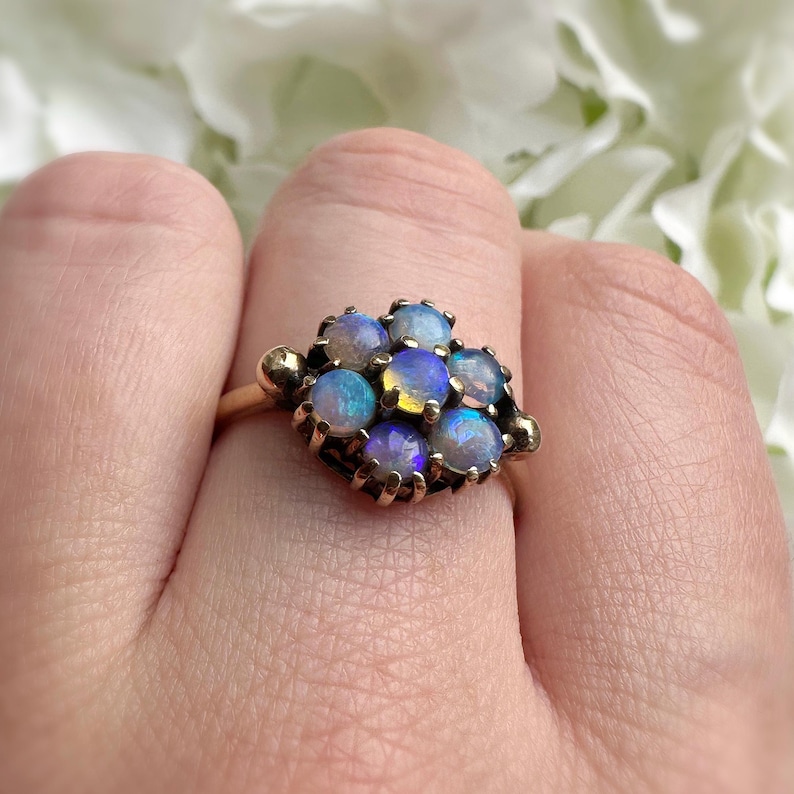 May include: A gold ring featuring a cluster of opals. The opals display a range of colors, including blue, purple, and yellow. The ring's band and setting are gold-toned, with a detailed design around each opal.