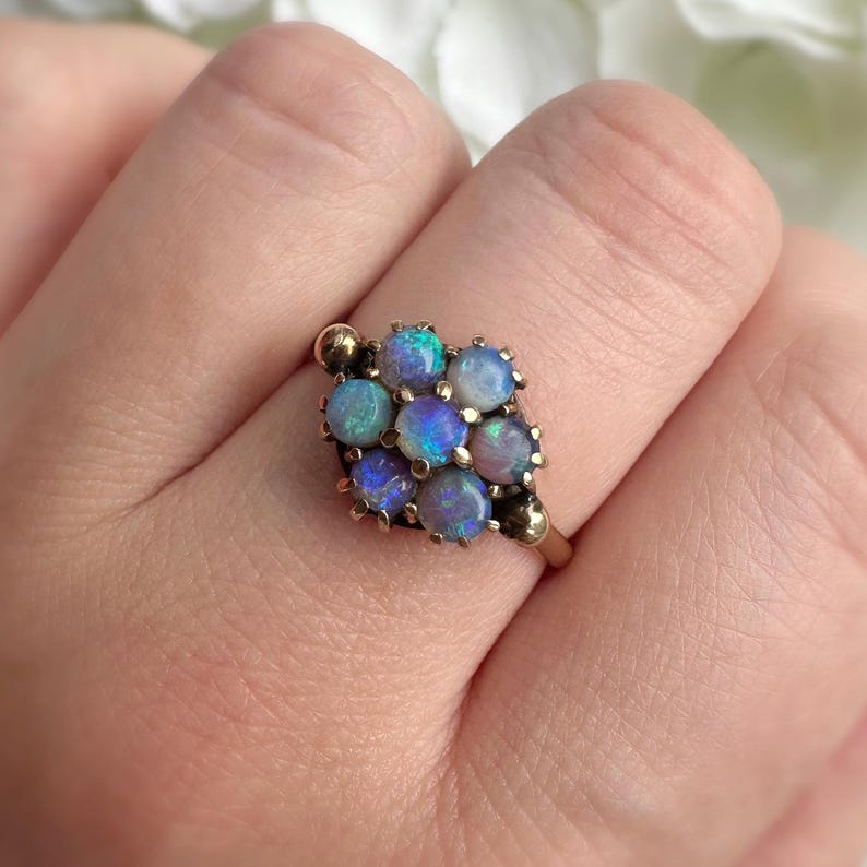 May include: A gold ring featuring a cluster of opals. The opals are a mix of blue, green, and purple hues. The ring has a classic design with a gold band and setting. The opals are arranged in a square shape.