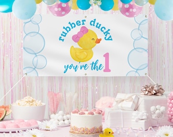 以下が含まれることがあります： 青い泡と「rubber ducky you're the 1」というピンクとブルーの文字が書かれた白いバナー。バナーの中央には、ピンクのリボンをつけた黄色のゴム製のオモチャの鴨がいる。バナーはピンクのテーブルクロスがかかったテーブルの上の紐から吊り下げられている。テーブルの上には、ピンクのフロスティングとスプリンクルがかけられた白いケーキと、黄色のゴム製のオモチャの鴨がある。