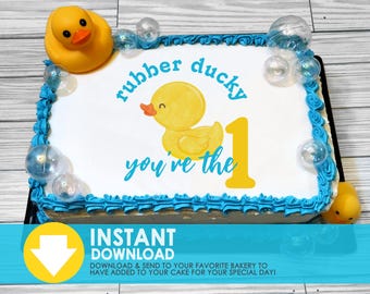 May include: A white cake with blue frosting and a yellow rubber ducky on top. The cake is decorated with the words "rubber ducky you're the 1".