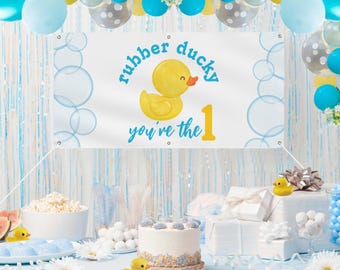 Puede incluir: Un pato de goma amarillo con una pancarta azul que dice "rubber ducky you're the 1". La pancarta está colgada de una cuerda azul y blanca. Hay globos azules y amarillos en el fondo.