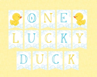 Puede incluir: Una pancarta blanca con letras azules y amarillas que deletrean "ONE LUCKY DUCK". La pancarta tiene un patrón de burbujas azules y blancas. Hay dos ilustraciones de patos de goma amarillos en la pancarta.
