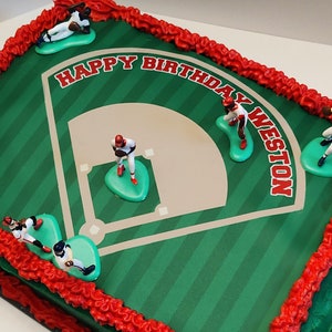 May include: A rectangular sheet cake decorated with a baseball field graphic and the text HAPPY BIRTHDAY WESTON. The cake features red frosting borders and five small plastic baseball player figurines positioned on the field surface.