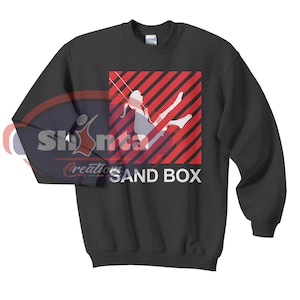 Peut inclure: Un sweat-shirt noir avec un motif à rayures rouges et blanches représentant une silhouette de personne sur une balançoire. Le texte "SAND BOX" est imprimé sous le motif.