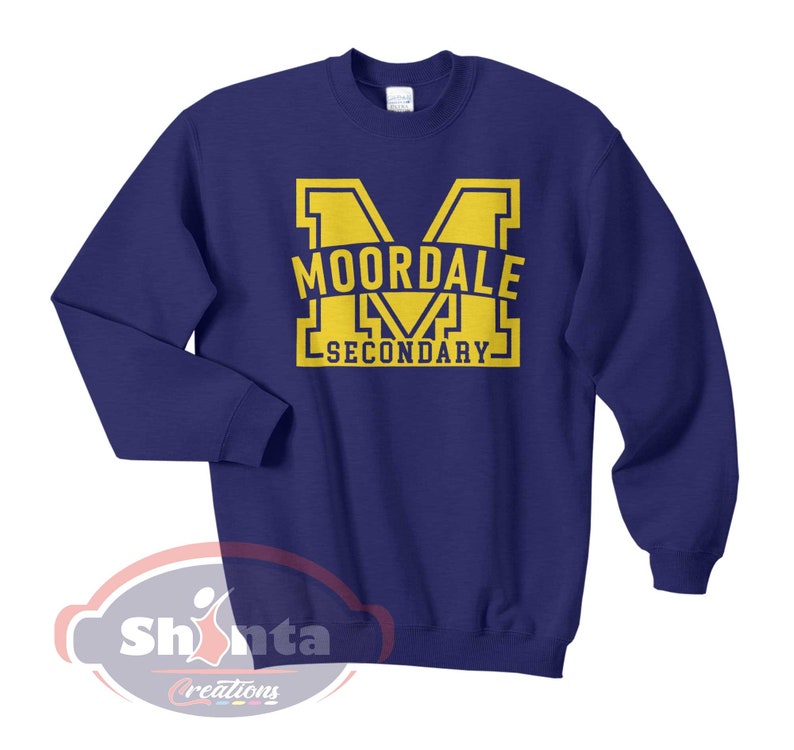 K&ouml;nnte beinhalten: Ein marineblauer Pullover mit einer gelben Grafik eines gro&szlig;en Buchstabens "M" mit dem Text "MOORDALE SECONDARY" im Buchstaben.