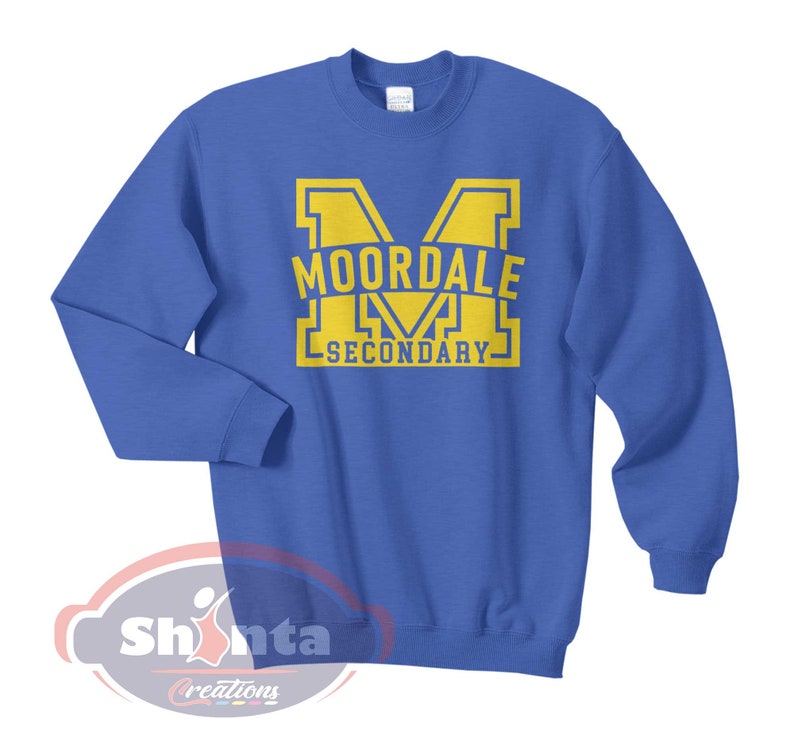 K&ouml;nnte beinhalten: Ein k&ouml;nigsblauer Pullover mit einer gelben Grafik eines gro&szlig;en Buchstabens "M" mit dem Text "MOORDALE SECONDARY" im Buchstaben.