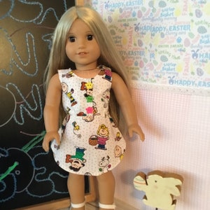 May include: A blonde doll wearing a white dress with a Peanuts character print. The dress has a pink polka dot pattern. The doll is wearing white shoes and is standing in front of a white background with a pink and white pattern. The background has the text "Happy Easter" repeated on it. There is a wooden Easter bunny decoration in the foreground.