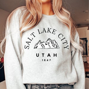 May include: A light gray sweatshirt with the text "SALT LAKE CITY UTAH 1847" in black, featuring a mountain range graphic. The sweatshirt is crew neck and long-sleeved.