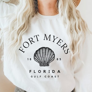 May include: White sweatshirt with a black graphic that reads "FORT MYERS" above a seashell illustration, with "1885" and "FLORIDA GULF COAST" below. The sweatshirt is a crew neck style.