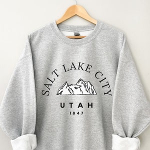 May include: A light gray sweatshirt with the text "SALT LAKE CITY UTAH 1847" in black, arched above a mountain graphic. The sleeves are cuffed, revealing a white interior. The sweatshirt is hanging on a wooden hanger.