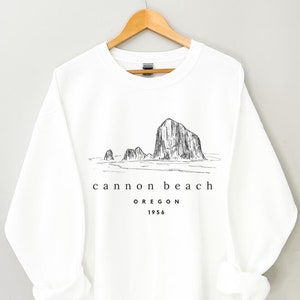 May include: White sweatshirt with a black and white illustration of Haystack Rock at Cannon Beach, Oregon. The text "cannon beach OREGON 1956" is printed below the illustration.