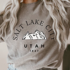 May include: A gray sweatshirt with the text "SALT LAKE CITY UTAH 1847" in black. Above the text is a white mountain range graphic. The sweatshirt is a crew neck style.