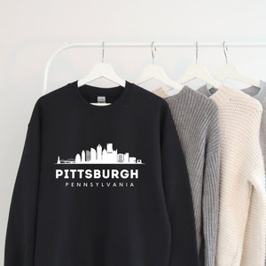 Może przedstawiać: Czarna bluza z białym nadrukiem panoramy Pittsburgha i tekstem "Pittsburgh, Pensylwania".