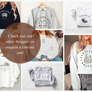 May include: A collage of images featuring various sweatshirts with state-themed designs. The sweatshirts are in different colors, including black, white, gray, and heather gray. The designs include a crab, mountains, a moose, cacti, and a mountain range. The text on the sweatshirts includes the names of states such as Maryland, Utah, Idaho, Arizona, and Alaska.