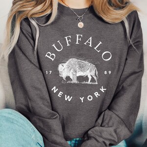 May include: A dark gray sweatshirt with a white graphic of a buffalo and the text "BUFFALO 1789 NEW YORK".