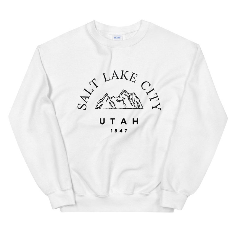 May include: White sweatshirt with a black graphic design. The design features the text "SALT LAKE CITY UTAH 1847" in a semi-circular arrangement around a mountain range illustration. The sweatshirt is a crew neck style.