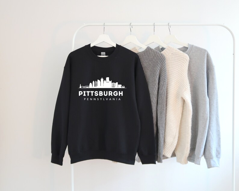 Może przedstawiać: Czarna bluza z białym nadrukiem panoramy Pittsburgha i tekstem "Pittsburgh, Pensylwania".