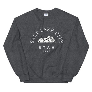 May include: Dark gray sweatshirt with the text "SALT LAKE CITY UTAH 1847" in white, arched above a mountain range graphic. The sweatshirt is a crew neck style.