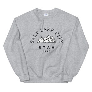 May include: A light gray sweatshirt with a crew neck. The front features a black graphic of mountains with the text "SALT LAKE CITY UTAH 1847" in a semi-circle above the mountains.