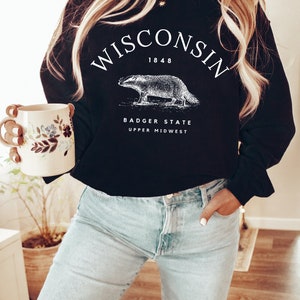 May include: Black sweatshirt with white text that reads "WISCONSIN 1848 BADGER STATE UPPER MIDWEST". A white illustration of a badger is centered on the sweatshirt.