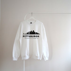 Może przedstawiać: Biała bluza z czarnym nadrukiem panoramy Pittsburgha i tekstem "Pittsburgh, Pensylwania".