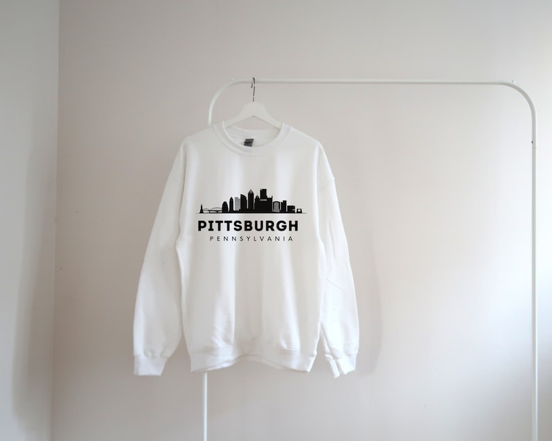 Może przedstawiać: Biała bluza z czarnym nadrukiem panoramy Pittsburgha i tekstem "Pittsburgh, Pensylwania".