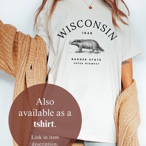 May include: A white t-shirt with a black graphic of a badger and the text "WISCONSIN 1848 BADGER STATE UPPER MIDWEST". The model is wearing the t-shirt with a brown cardigan and blue jeans.