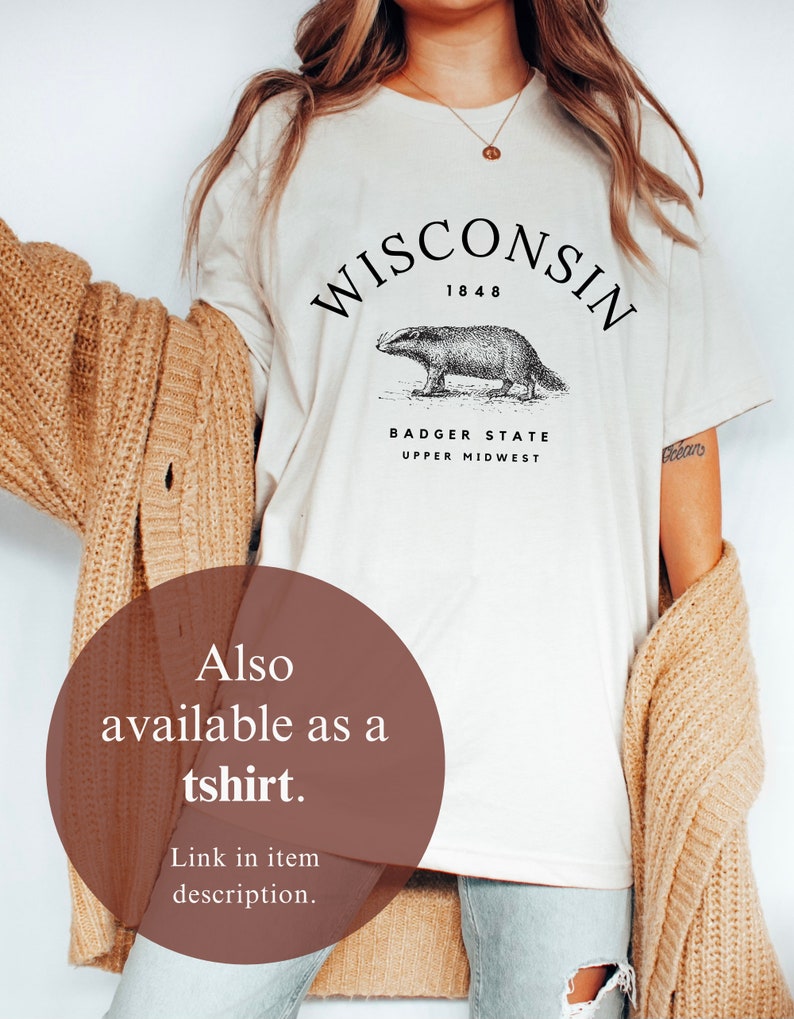 May include: A white t-shirt with a black graphic of a badger and the text "WISCONSIN 1848 BADGER STATE UPPER MIDWEST". The model is wearing the t-shirt with a brown cardigan and blue jeans.
