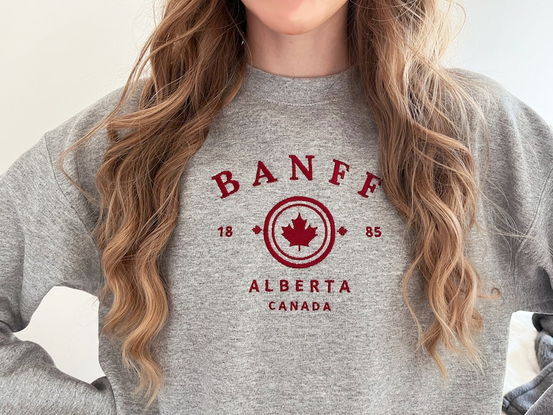 Puede incluir: Sudadera gris con un estampado gr&aacute;fico rojo que dice "BANFF 18 85 ALBERTA CANADA" con una hoja de arce roja dentro de un c&iacute;rculo.