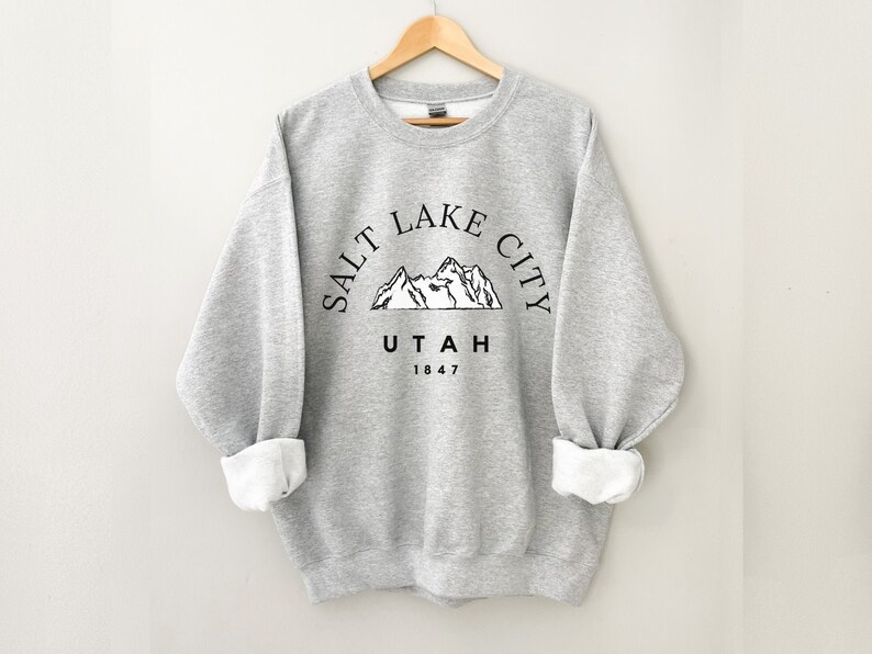 May include: A light gray sweatshirt with the text "SALT LAKE CITY UTAH 1847" in black, arched above a mountain graphic. The sleeves are cuffed, revealing a white interior. The sweatshirt is hanging on a wooden hanger.