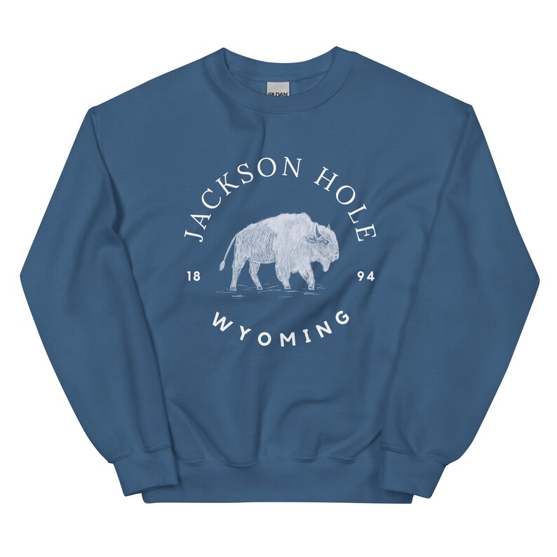 以下が含まれることがあります： 白い「Jackson Hole 1894 Wyoming」の文字が書かれた円の中に、白いバイソンのグラフィックが描かれた青いスウェットシャツです。