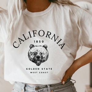 May include: White t-shirt with a black graphic design featuring a bear and the text "CALIFORNIA 1850 GOLDEN STATE WEST COAST".