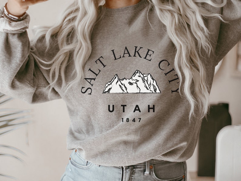 May include: A gray sweatshirt with the text "SALT LAKE CITY UTAH 1847" in black. Above the text is a white mountain range graphic. The sweatshirt is a crew neck style.