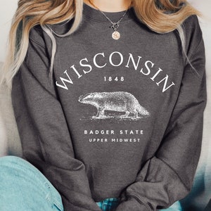 May include: A dark gray sweatshirt with a white graphic design featuring the text "WISCONSIN 1848" and a badger illustration. The text "BADGER STATE UPPER MIDWEST" is also included in the design.