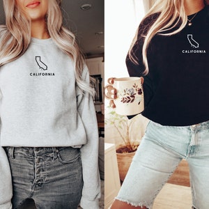 May include: Two women wearing sweatshirts with a California outline and the word "California" printed on them. One sweatshirt is grey and the other is black.