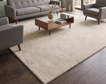 May include: A large, rectangular, solid beige area rug with a subtle, low-pile texture. The rug is positioned on a dark wood floor in a living room setting, centered beneath a wooden coffee table with a glass top and two gray upholstered sofas.